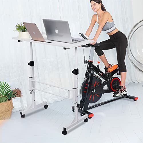 Woman using an exercise bike with a desk setup, two laptops on the desk.
